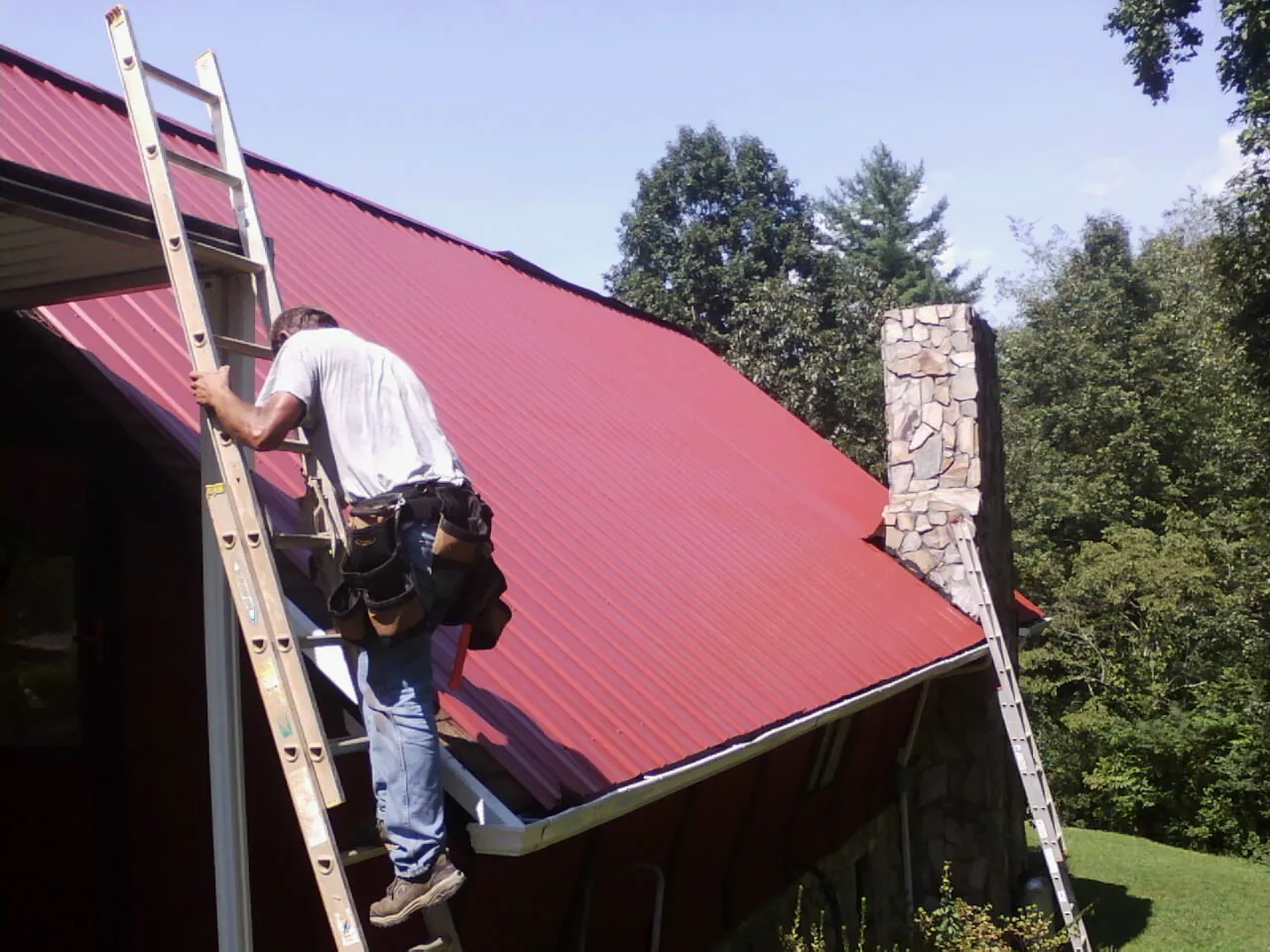 Roofer on a ladder beside a red metal roof and stone chimney — Soffit & Fascia Repair by Roofing Brothers in Wyndmere